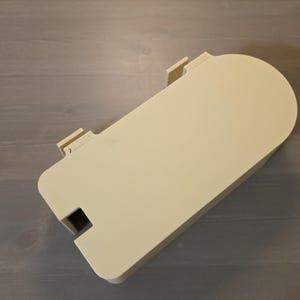 May include: A cream-colored, rectangular plastic cover with one rounded end. The object features two small, protruding rectangular tabs along one of the straight edges and a small, square-shaped cutout near the opposite corner of the same side.