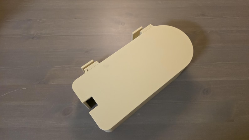 May include: A cream-colored, rectangular plastic cover with one rounded end. The object features two small, protruding rectangular tabs along one of the straight edges and a small, square-shaped cutout near the opposite corner of the same side.
