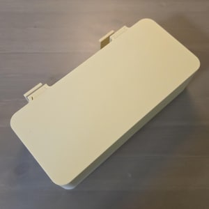 May include: A rectangular, light cream-colored plastic storage box with rounded corners and a flat, solid lid. The container features two small, protruding hinge-like tabs along one of the longer sides, indicating a hinged opening mechanism.