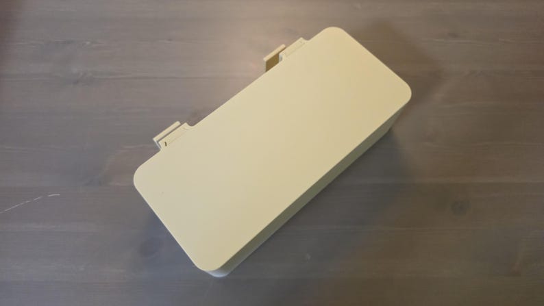 May include: A rectangular, light cream-colored plastic storage box with rounded corners and a flat, solid lid. The container features two small, protruding hinge-like tabs along one of the longer sides, indicating a hinged opening mechanism.