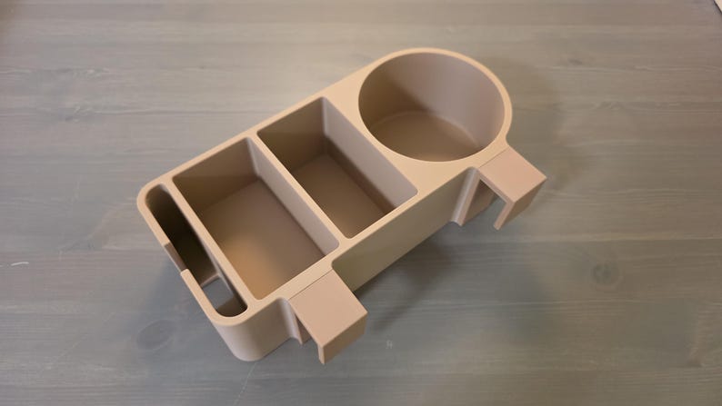 May include: A beige plastic desk organizer featuring a circular cup holder on one side, two rectangular storage compartments in the center, and a narrow vertical slot on the opposite end, all integrated into a single, unified structure.