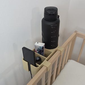 May include: A beige plastic bedside organizer with multiple compartments attached to the wooden railing of a crib. The organizer holds a black smartphone, a small rectangular box with a floral pattern, and a large matte black water bottle.