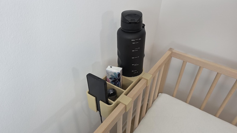 May include: A beige plastic bedside organizer with multiple compartments attached to the wooden railing of a crib. The organizer holds a black smartphone, a small rectangular box with a floral pattern, and a large matte black water bottle.