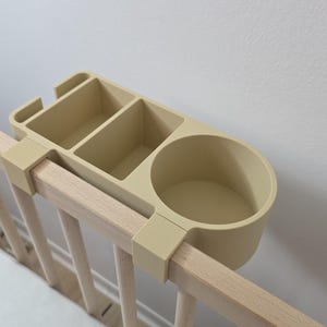 May include: A beige plastic bedside organizer with a circular cup holder and two rectangular compartments, designed to clip onto the top rail of a wooden crib. The organizer features a smooth, matte finish and a functional, multi-section design.