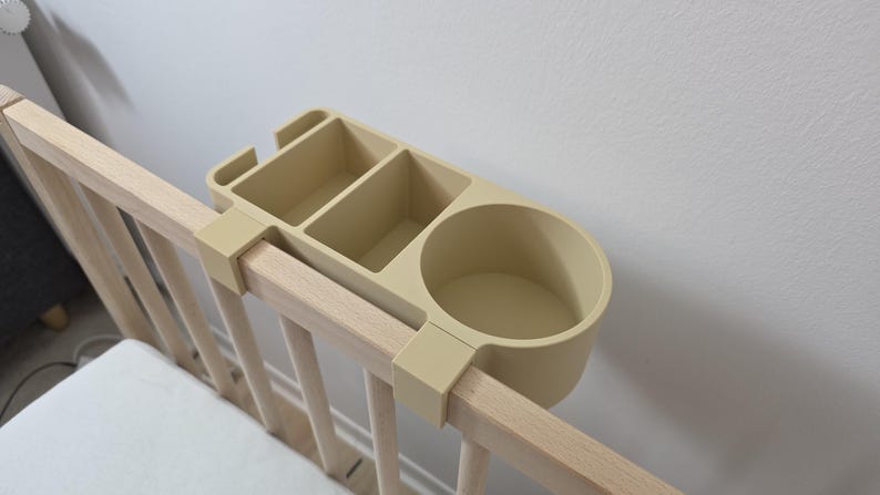 May include: A beige plastic bedside organizer with a circular cup holder and two rectangular compartments, designed to clip onto the top rail of a wooden crib. The organizer features a smooth, matte finish and a functional, multi-section design.