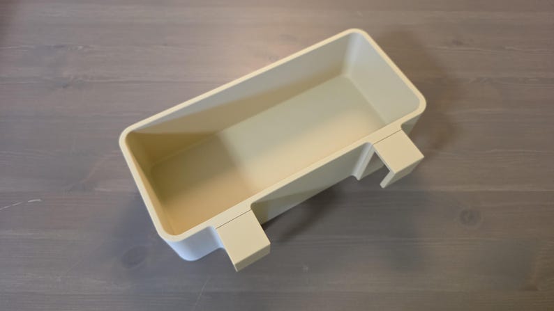 May include: A rectangular, off-white plastic storage bin featuring a hollow interior and two integrated hooks on one side, designed for hanging or mounting. The container has smooth, rounded corners and a solid, uniform matte finish throughout.