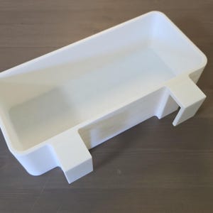 May include: A rectangular white plastic storage bin featuring two integrated hooks on one side, designed to hang over an edge. The container has a smooth, matte finish with rounded corners and a deep, open interior for holding various small items.