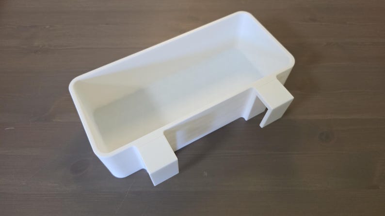 May include: A rectangular white plastic storage bin featuring two integrated hooks on one side, designed to hang over an edge. The container has a smooth, matte finish with rounded corners and a deep, open interior for holding various small items.
