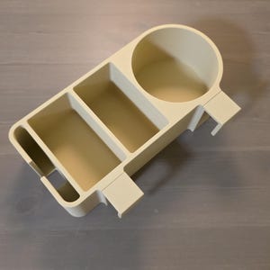 May include: A beige plastic desk organizer featuring a circular cup holder, two rectangular compartments, and a narrow slot on the side. The organizer has a flat base and integrated side clips designed for attachment to a flat edge or surface.
