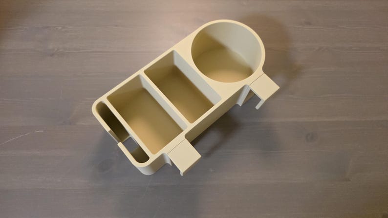 May include: A beige plastic desk organizer featuring a circular cup holder, two rectangular compartments, and a narrow slot on the side. The organizer has a flat base and integrated side clips designed for attachment to a flat edge or surface.