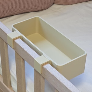 May include: A rectangular, cream-colored plastic storage bin featuring two integrated hooks designed to hang securely over the top rail of a wooden bed frame. The container has smooth, rounded corners and an open top for accessible storage.