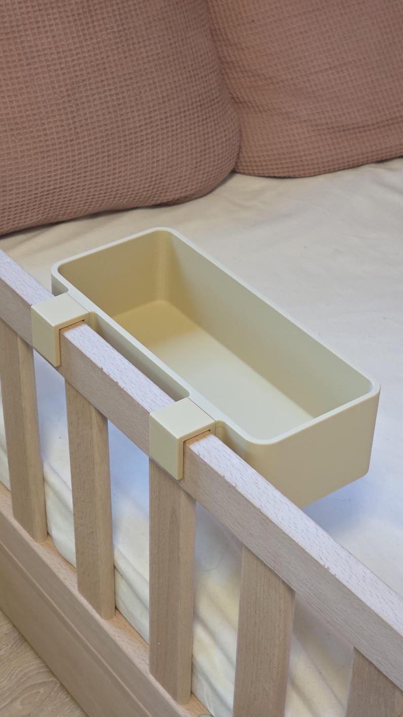 May include: A rectangular, cream-colored plastic storage bin featuring two integrated hooks designed to hang securely over the top rail of a wooden bed frame. The container has smooth, rounded corners and an open top for accessible storage.