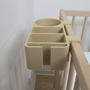 May include: A beige plastic bedside organizer with a circular cup holder and three rectangular compartments, featuring a hook mechanism that attaches securely to the side rail of a light-colored wooden crib for convenient storage access.