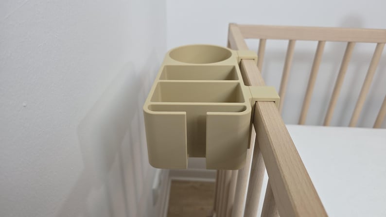 May include: A beige plastic bedside organizer with a circular cup holder and three rectangular compartments, featuring a hook mechanism that attaches securely to the side rail of a light-colored wooden crib for convenient storage access.