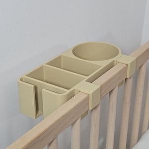 Könnte beinhalten: Ein beigefarbener Babybett-Organizer aus Kunststoff mit einem Becherhalter und zwei Fächern, der an einer Holz-Bettseite befestigt ist. Der Organizer ist für Babyutensilien konzipiert.