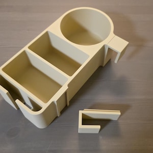 May include: A beige plastic desk organizer featuring a circular cup holder, two rectangular storage compartments, and a side slot. The organizer includes a separate, detachable U-shaped mounting bracket for attachment to a flat surface edge.