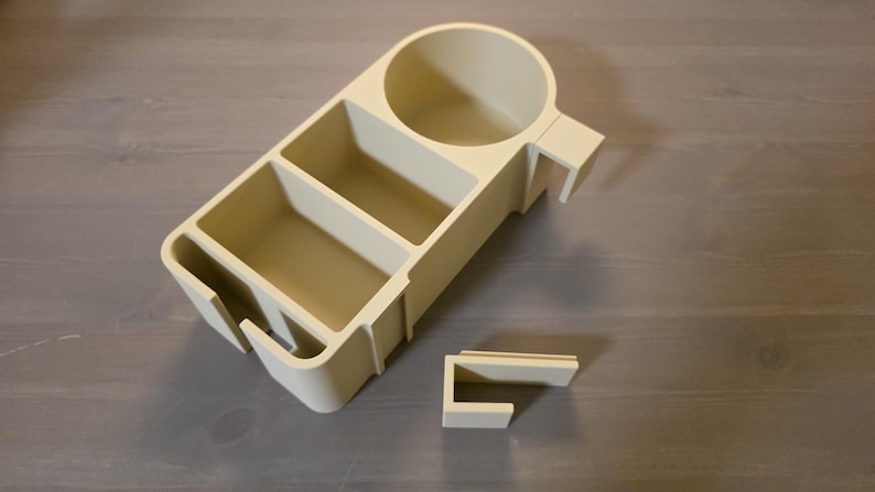May include: A beige plastic desk organizer featuring a circular cup holder, two rectangular storage compartments, and a side slot. The organizer includes a separate, detachable U-shaped mounting bracket for attachment to a flat surface edge.