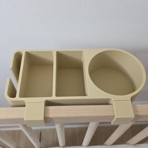 May include: A beige plastic bedside organizer featuring a circular cup holder, two rectangular compartments, and a narrow slot on the side. The organizer is designed with two hooks that attach securely over the top rail of a wooden crib frame.