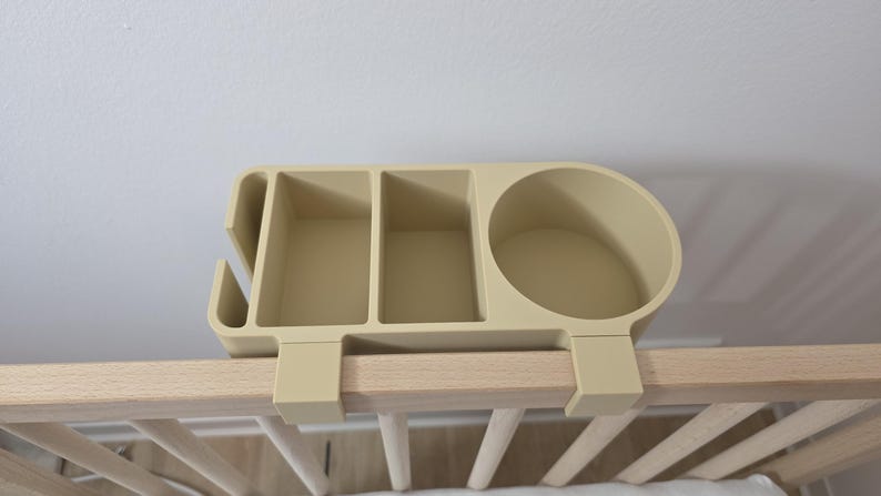 May include: A beige plastic bedside organizer featuring a circular cup holder, two rectangular compartments, and a narrow slot on the side. The organizer is designed with two hooks that attach securely over the top rail of a wooden crib frame.