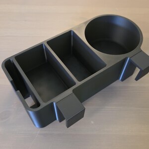 May include: A matte black plastic organizer tray featuring a circular cup holder, two rectangular storage compartments, and a narrow slot. The unit includes two protruding rectangular hooks on the side for mounting purposes on a flat edge.