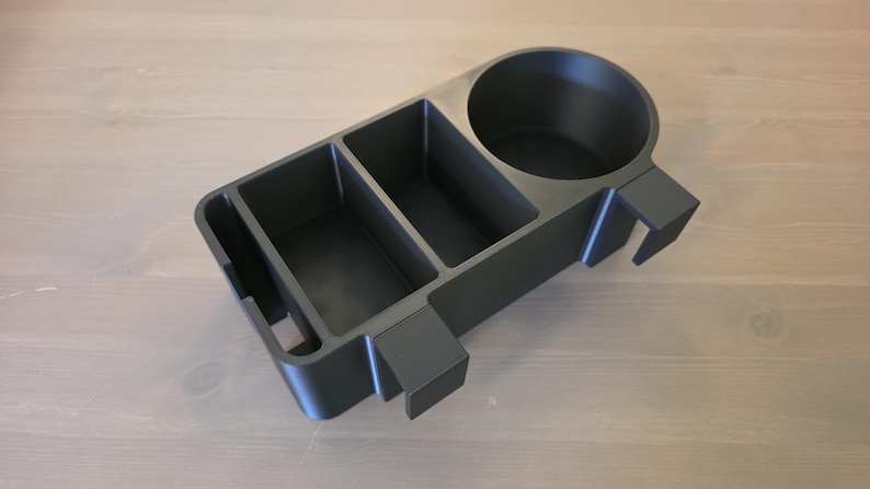 May include: A matte black plastic organizer tray featuring a circular cup holder, two rectangular storage compartments, and a narrow slot. The unit includes two protruding rectangular hooks on the side for mounting purposes on a flat edge.