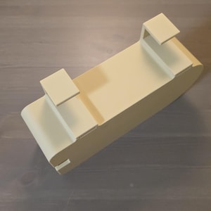 May include: A cream-colored, rectangular over-the-door storage organizer featuring two integrated hooks on the top edge. The unit has a smooth, solid finish with rounded corners and a deep, open-top compartment designed for hanging storage.