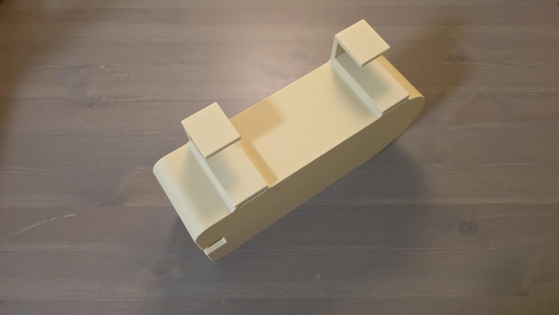 May include: A cream-colored, rectangular over-the-door storage organizer featuring two integrated hooks on the top edge. The unit has a smooth, solid finish with rounded corners and a deep, open-top compartment designed for hanging storage.
