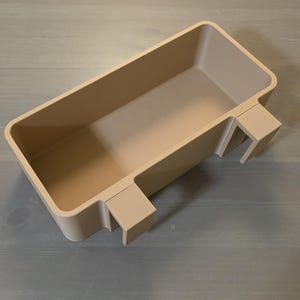 May include: A rectangular beige storage bin with a smooth, matte finish. The container features two integrated hooks on one side, designed to hang over an edge. The bin has a deep, open interior and rounded corners, providing a simple, functional design.