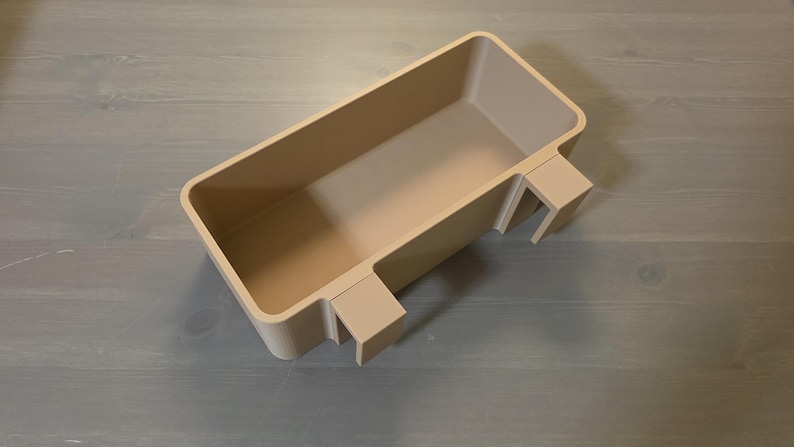 May include: A rectangular beige storage bin with a smooth, matte finish. The container features two integrated hooks on one side, designed to hang over an edge. The bin has a deep, open interior and rounded corners, providing a simple, functional design.