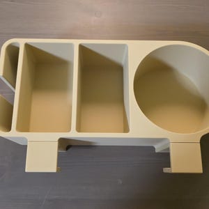 May include: A beige desk organizer featuring a circular cup holder on the right side, two rectangular compartments in the center, and a narrow vertical slot on the left side. The unit includes two downward-facing clips for mounting purposes.