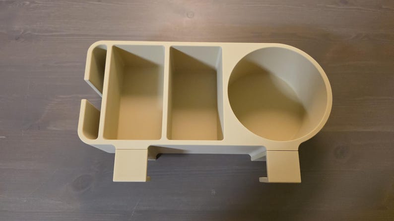 May include: A beige desk organizer featuring a circular cup holder on the right side, two rectangular compartments in the center, and a narrow vertical slot on the left side. The unit includes two downward-facing clips for mounting purposes.