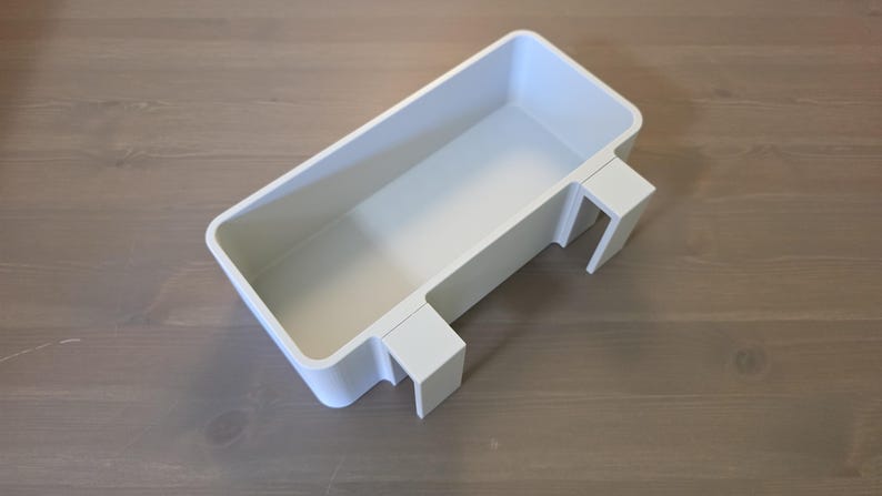 May include: A rectangular white plastic storage bin featuring an open top and two integrated hooks on one side, designed to hang over the edge of a flat surface. The container has smooth, rounded corners and a clean, solid white finish throughout.