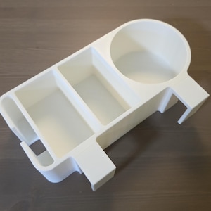 May include: A white plastic desk organizer featuring a circular cup holder on one side, two rectangular compartments in the center, and a narrow slot on the opposite end. The organizer has a flat base and integrated side clips for mounting.