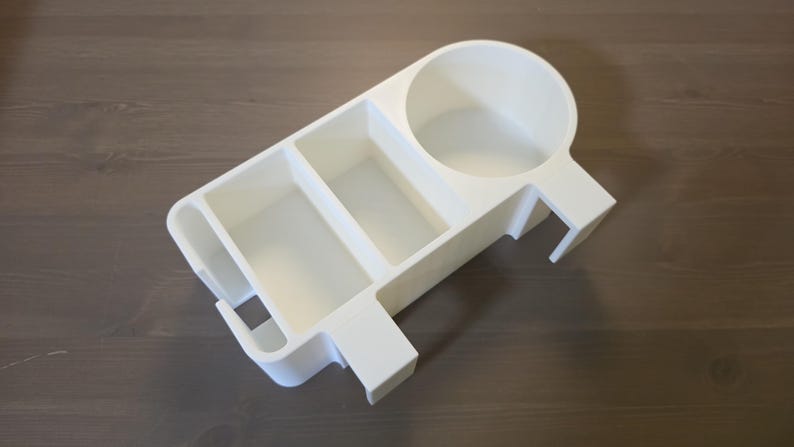May include: A white plastic desk organizer featuring a circular cup holder on one side, two rectangular compartments in the center, and a narrow slot on the opposite end. The organizer has a flat base and integrated side clips for mounting.