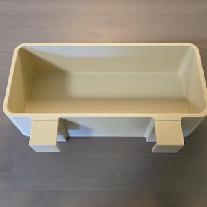 May include: A rectangular, cream-colored plastic storage bin featuring an open top and two integrated hooks on the front side, designed for hanging or mounting. The container has smooth, rounded corners and a solid, uniform matte finish throughout.