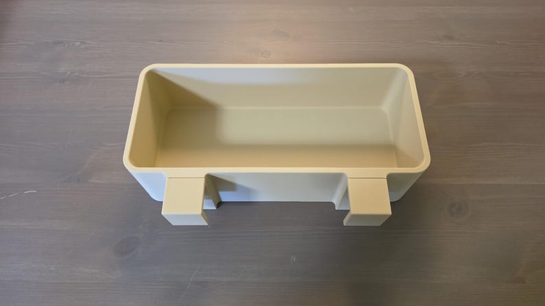 May include: A rectangular, cream-colored plastic storage bin featuring an open top and two integrated hooks on the front side, designed for hanging or mounting. The container has smooth, rounded corners and a solid, uniform matte finish throughout.