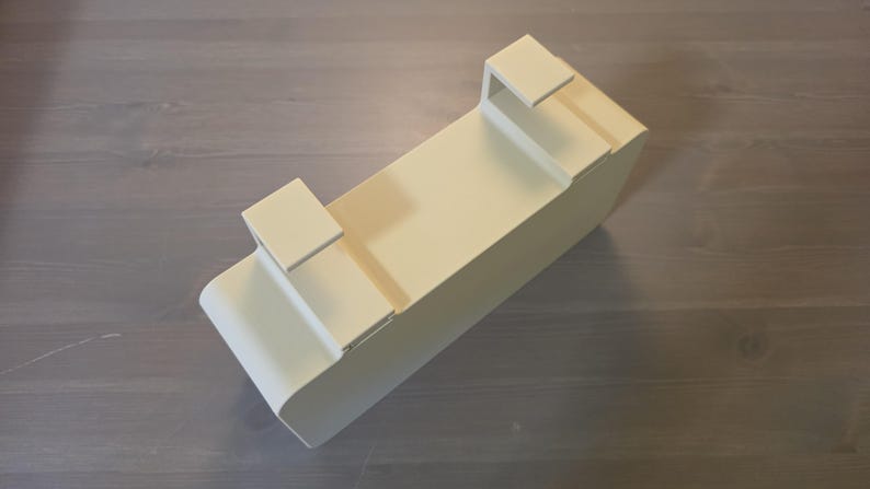 May include: A rectangular, off-white metal storage bin featuring two integrated hooks on the top edge. The container has a smooth, solid finish with rounded corners on one side and a flat, open-top design, constructed for hanging or mounting purposes.