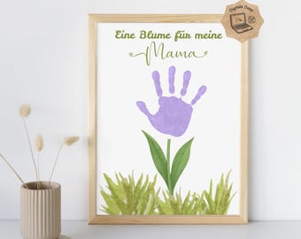 May include: A framed print with a handprint flower design. The flower is made from a purple handprint, with green leaves and grass. The text "Eine Blume für meine Mama" is written above the flower in green script. A small digital file icon is in the top right corner.