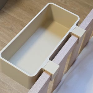 May include: A rectangular, cream-colored plastic storage bin featuring a hook-style attachment mechanism. The bin is securely mounted onto the vertical wooden slats of a bed frame, providing an open, accessible compartment for holding small items.