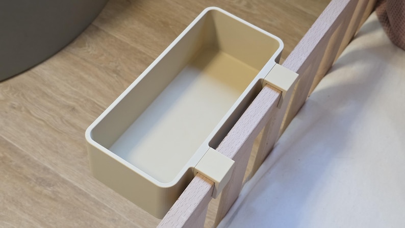 May include: A rectangular, cream-colored plastic storage bin featuring a hook-style attachment mechanism. The bin is securely mounted onto the vertical wooden slats of a bed frame, providing an open, accessible compartment for holding small items.
