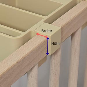 May include: A beige plastic storage organizer with multiple compartments attached to a light-colored wooden crib rail. The organizer features a mounting bracket with red and blue arrows indicating the width and height measurements labeled as Breite and H&ouml;he.