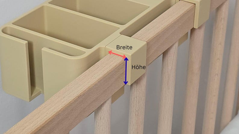 May include: A beige plastic storage organizer with multiple compartments attached to a light-colored wooden crib rail. The organizer features a mounting bracket with red and blue arrows indicating the width and height measurements labeled as Breite and H&ouml;he.