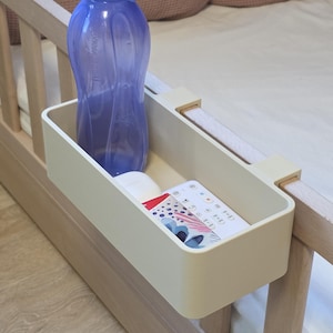 May include: A beige rectangular bedside storage caddy with two hooks attached to a wooden bed frame. Inside the caddy sits a translucent purple plastic water bottle and a white remote control with colorful circular icons and text printed on its surface.