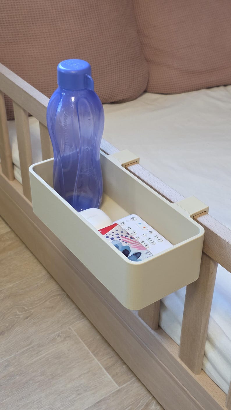 May include: A beige rectangular bedside storage caddy with two hooks attached to a wooden bed frame. Inside the caddy sits a translucent purple plastic water bottle and a white remote control with colorful circular icons and text printed on its surface.