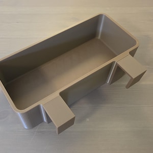 May include: A rectangular, taupe-colored plastic storage bin featuring two integrated hooks on one side, designed for hanging. The container has a smooth, matte finish with rounded corners and a deep, open interior space for holding various items.