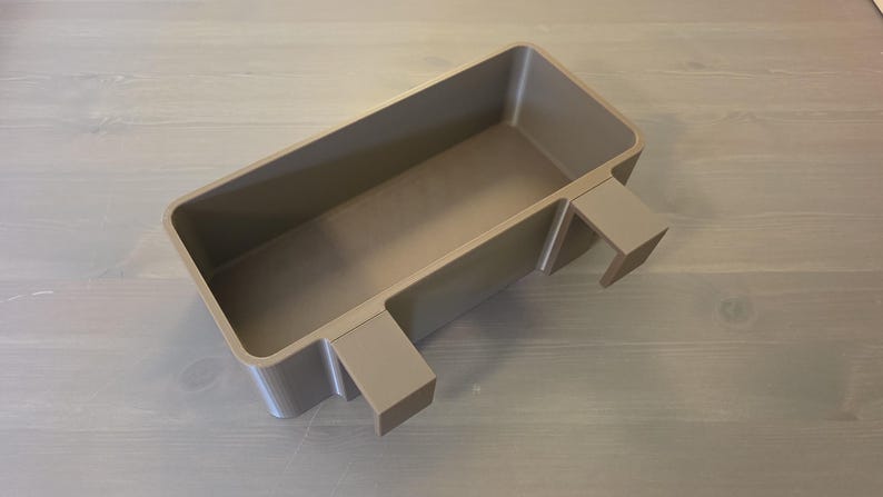 May include: A rectangular, taupe-colored plastic storage bin featuring two integrated hooks on one side, designed for hanging. The container has a smooth, matte finish with rounded corners and a deep, open interior space for holding various items.