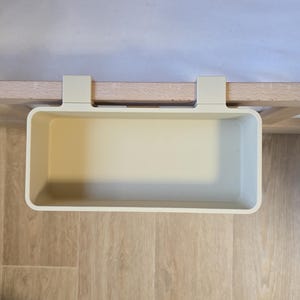 May include: A rectangular, off-white plastic storage bin featuring two integrated hooks designed to hang over the edge of a flat surface. The container has a smooth, solid-colored interior and exterior with rounded corners and a wide, open top.