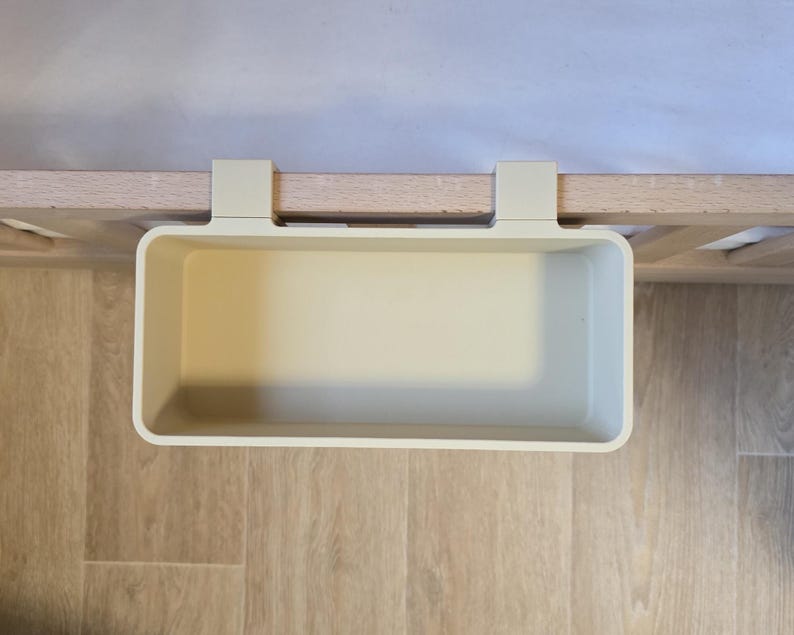 May include: A rectangular, off-white plastic storage bin featuring two integrated hooks designed to hang over the edge of a flat surface. The container has a smooth, solid-colored interior and exterior with rounded corners and a wide, open top.