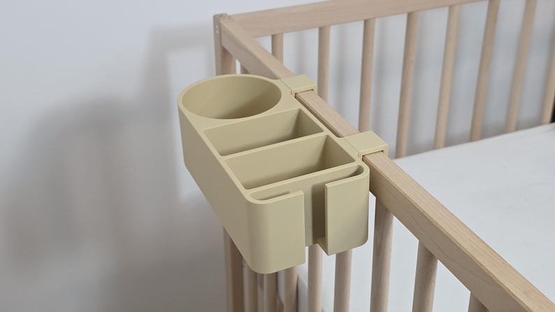May include: A beige plastic bedside organizer with a circular cup holder, two rectangular compartments, and a side slot, designed to attach securely to the wooden railing of a crib. The organizer features a smooth, matte finish and a functional design.