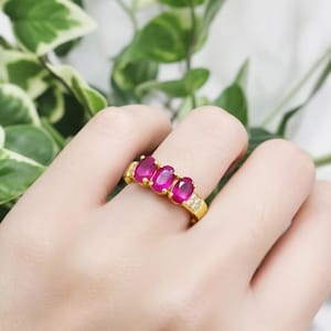 May include: A gold ring featuring three oval-cut, vibrant red gemstones. The ring is accented with small, clear stones on the sides. The ring is a piece of jewelry.